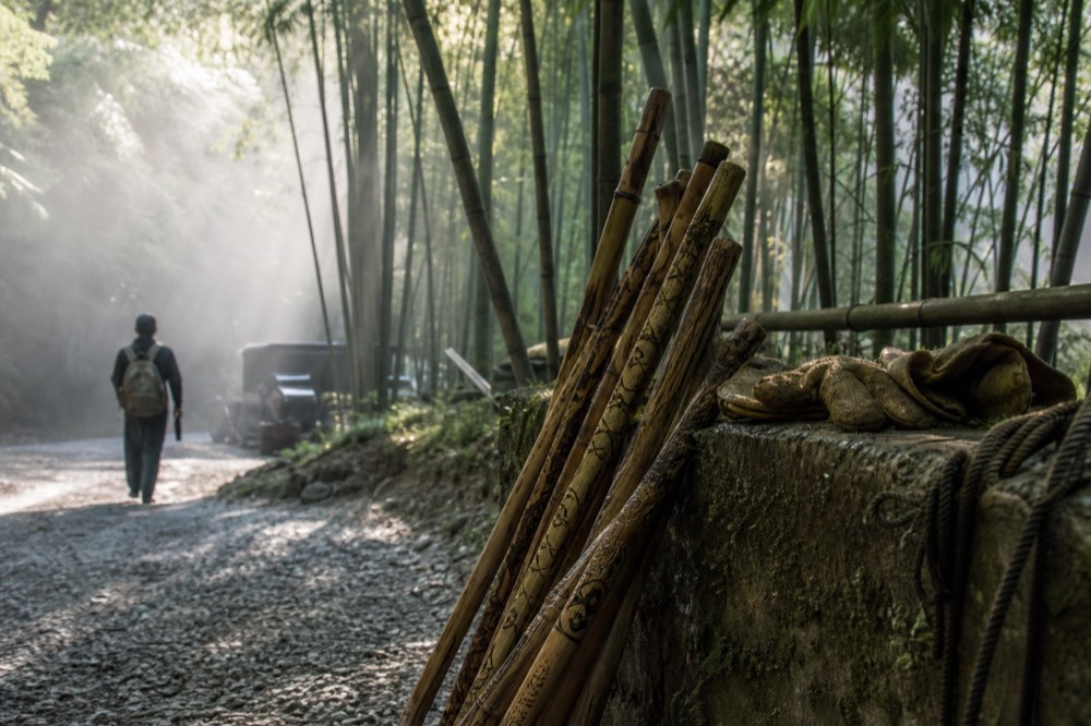 A cluster of carved wooden walking sticks leaning against a low wall beside worn canvas gloves and a coiled rope, early sun raking across the gravel