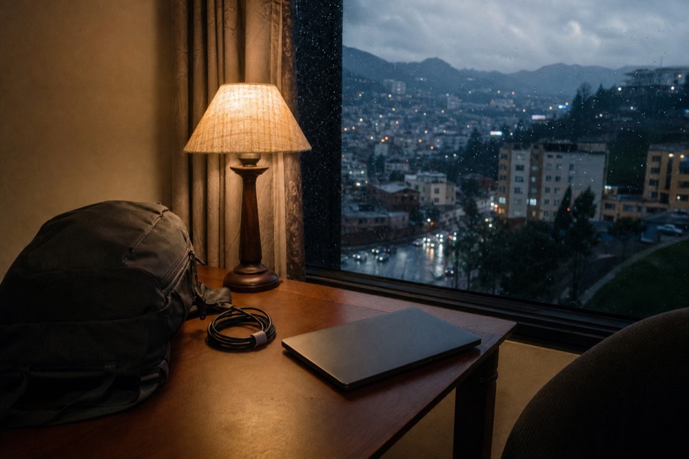 A closed laptop on a hotel desk beside a daypack and a coiled charging cable, warm lamplight, dark window and city glow behind