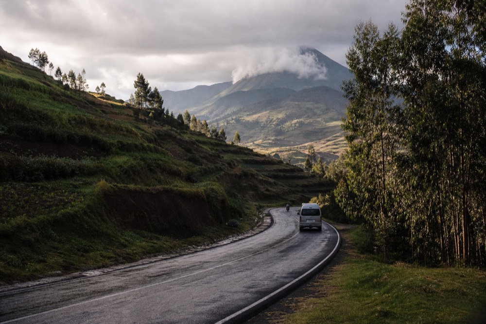 A rain-slicked tarmac highway climbing through steep green terraced hillsides toward volcanic peaks under a heavy grey sky, roadside eucalyptus trees