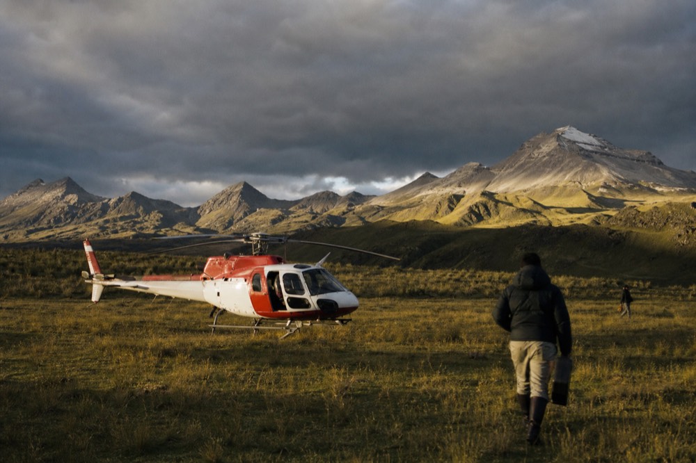A small red and white helicopter sitting on a grass airstrip with rotor blades still, long shadows across the field, a line of volcanic peaks behind