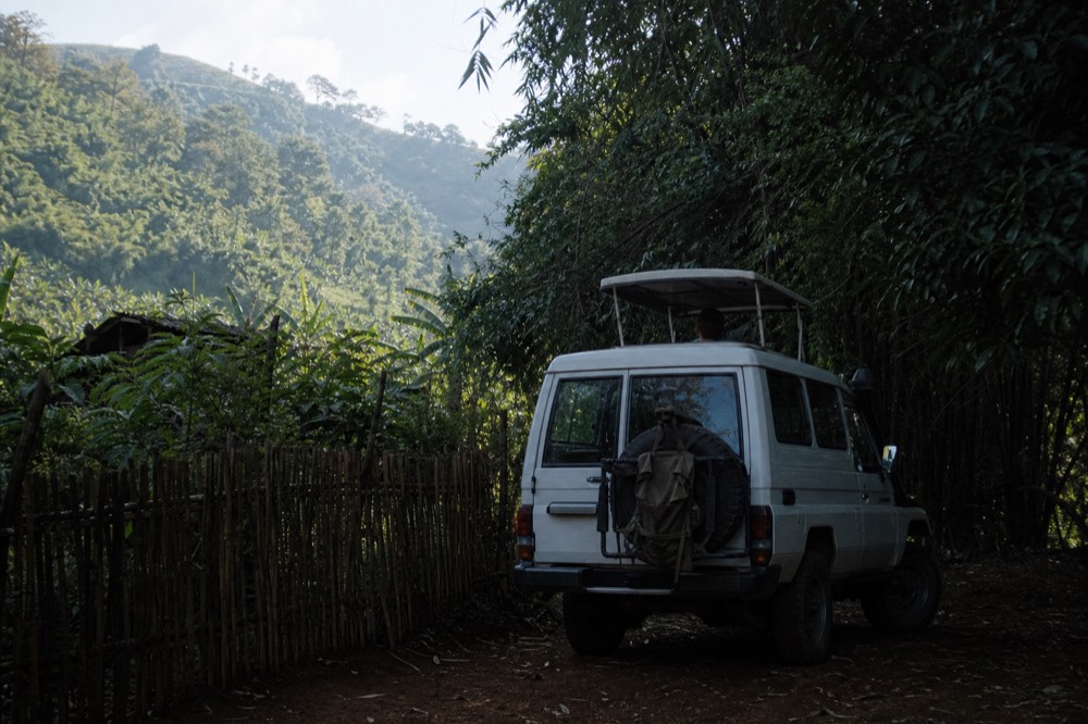 A white Toyota Land Cruiser with the roof hatch open parked on red earth beside a bamboo fence, luggage strapped to the rear, hills fading behind