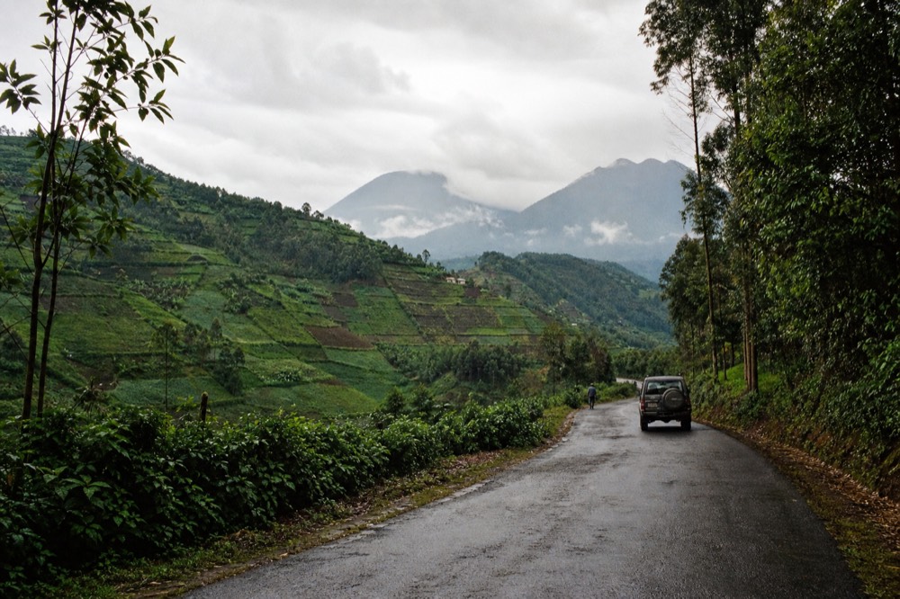 A tarmac road curving through steep terraced hillsides of green cropland in northern Rwanda, volcano silhouettes rising in the far distance under high cloud