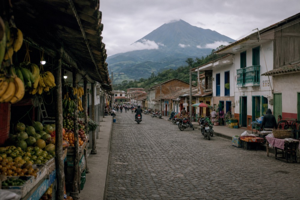 A busy small-town market street with produce stalls, parked motorbikes and painted shopfronts under an overcast sky, green hills closing the far end